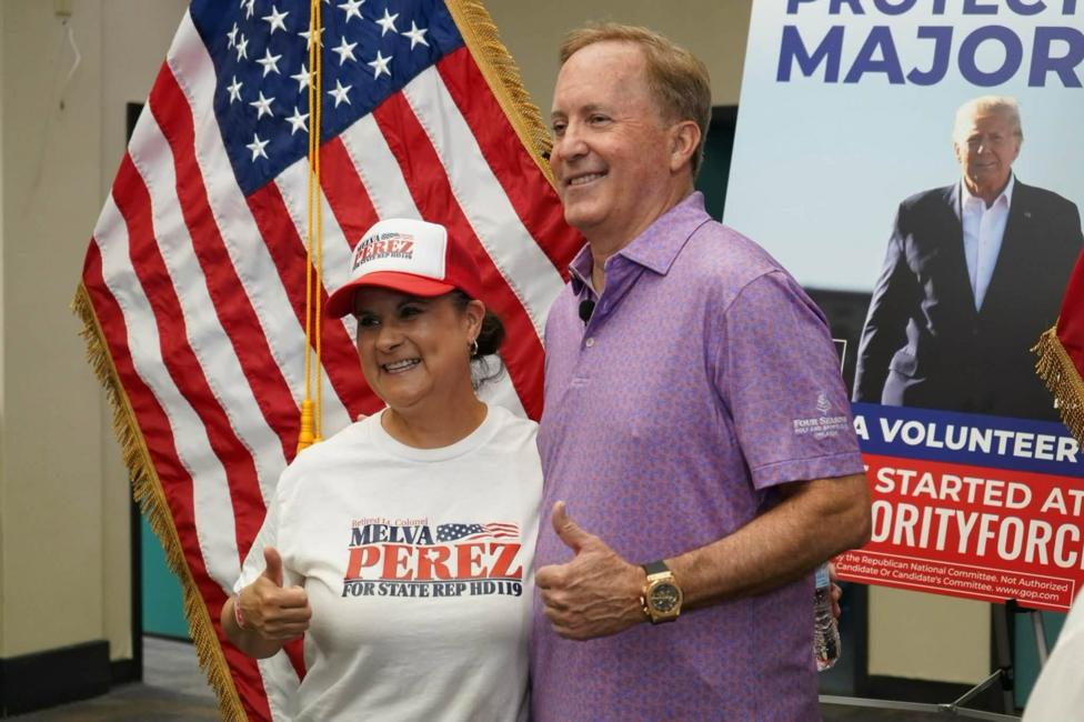 Ken Paxton and a female supporter pose in front of a large US flag and a campaign poster featuring Donald Trump. The woman wears a white campaign T-shirt and red-and-white baseball cap. Paxton wears a purple polo shirt. Both give a thumbs-up gesture.