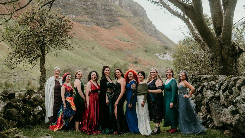 Eleven women wearing colourful dresses stand between two dry stone walls in a rural valley. Trees frame the group on either side, while a steep rocky hillside rises in the background.