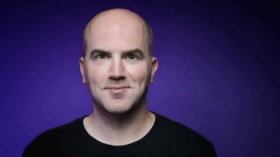 Composer Sam Watts looks directly into the camera. He is standing against a plain purple background.