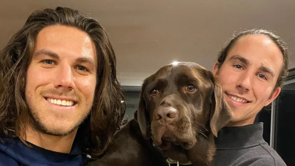 A young man with shoulder-length brown hair and light stubble smiling at the camera, and another young man to his right and a brown dog in between them