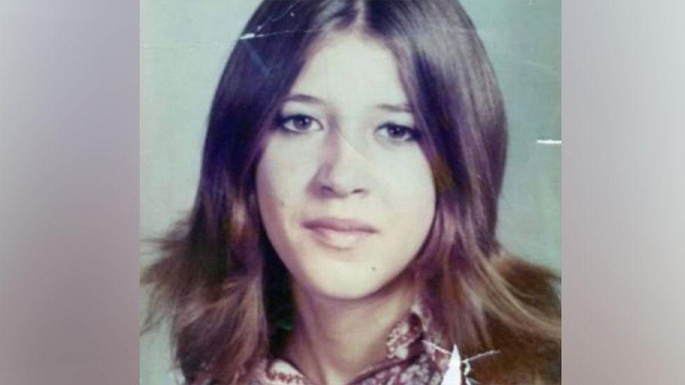 Лора Эйм на школьной фотографии 1970-х годов. У неё длинные каштановые волосы, разделённые на прямой пробор, карие глаза, на ней блузка с узором.