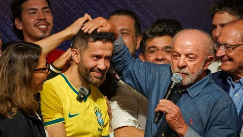 André Janones, wearing a yellow Brazilian football shirt, smiles as President Lula puts his right hand on top of his head in front of other jubilant supporters. Lula is wearing a blue denim shirt and holds a microphone in his left hand, which has a missing little finger.