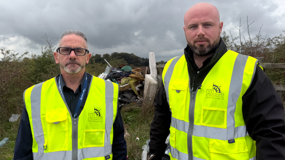 Anthony Horan and Ashley Hanley are both wearing yellow high-vis jackets. They are standing in front of a mound of fly-tipped waste including a mattress.