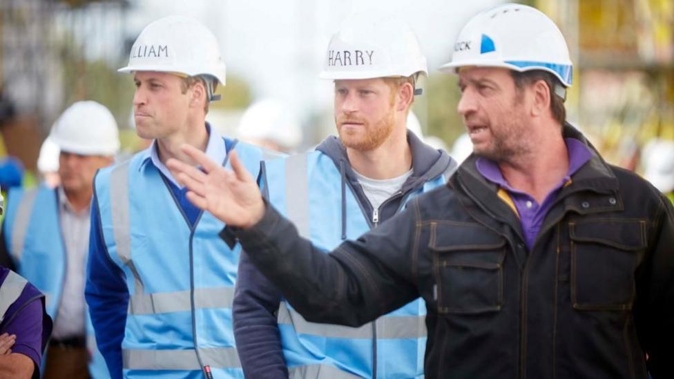 Prince William, Prince Harry and Nick Knowles, all in hard hats as Knowles talks and gestures to something out of frame