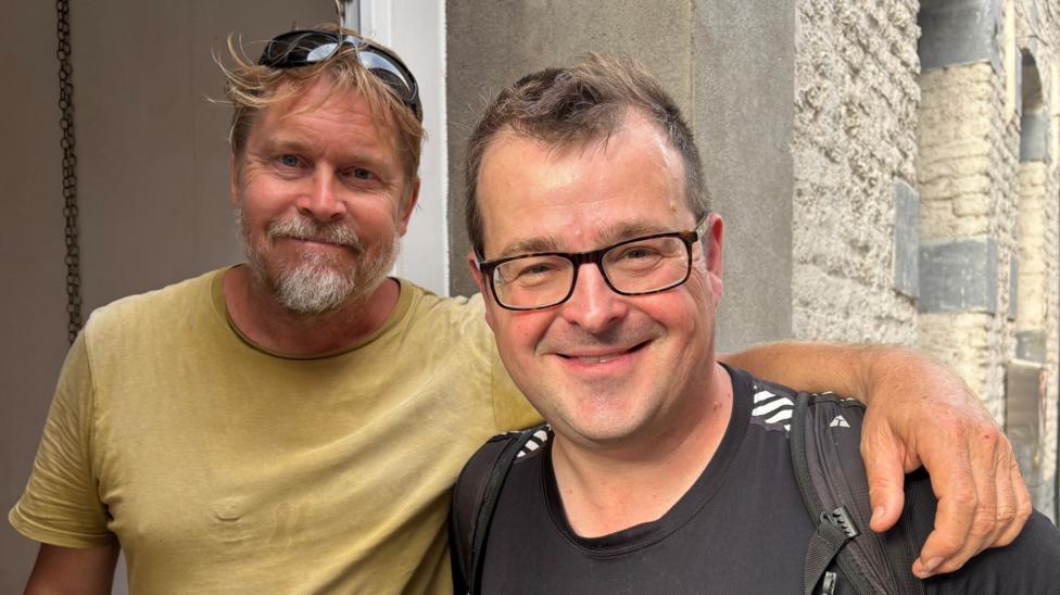 Global sarguzaştçi Karl Buşby BBC muxbiri Kevin Şusmitning yelkasiga qölini qöyib turibdi.