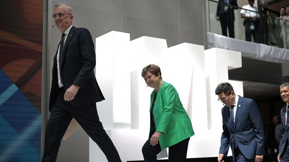 Three people walking up stairs one behind each other against a backdrop of white letters spelling IMF. 