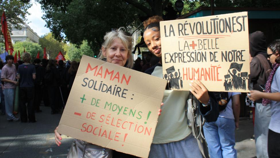 A young woman and her mother (to the left) stand together holding placards in French