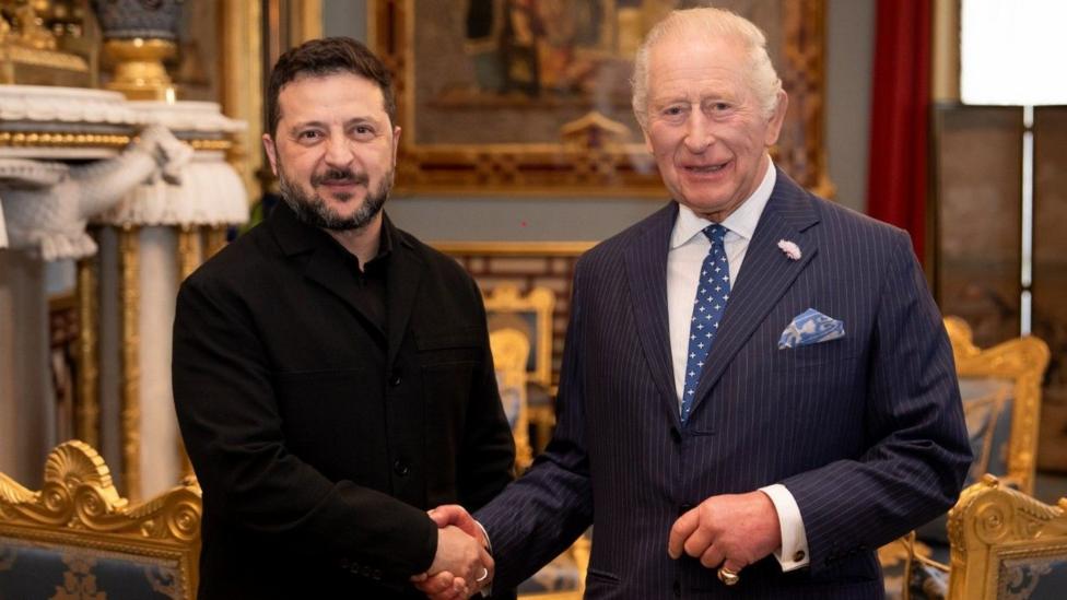 Volodymyr Zelensky and King Charles look towards the camera while smiling and shaking hands.