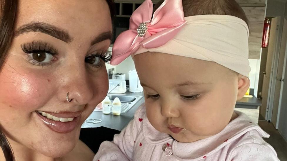 Sophie Price (left) is a young woman who has brown eyes and brown hair. She is looking at the camera and smiling. She is holding her daughter Florence (right) who is a baby. She wears a pink headband which has a large pink bow on it. She is looking down away from the camera.