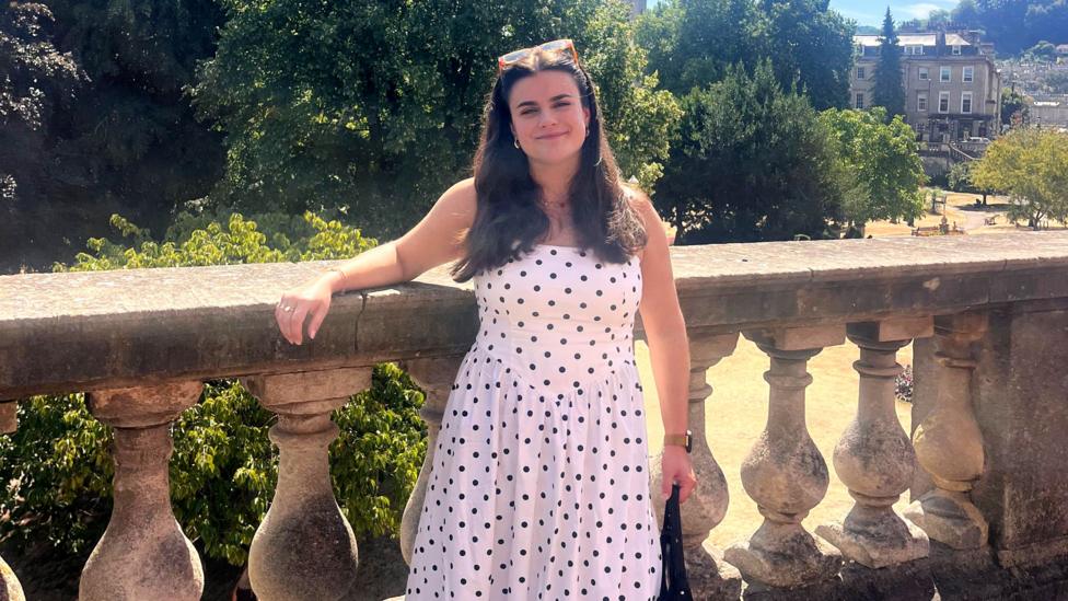 Uzun qora soçli yoş ayol Ella Kipling kameraga tabassum qilmoqda. U qora nuqtali uzun oq köylak kiygan va orqasida qadimiy binolar körinib turgan katta park oldida turibdi.