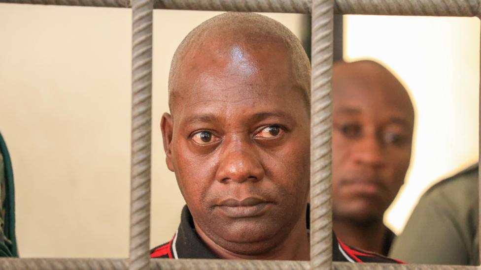 A close shot of a bald man looking through the bars of a cell
