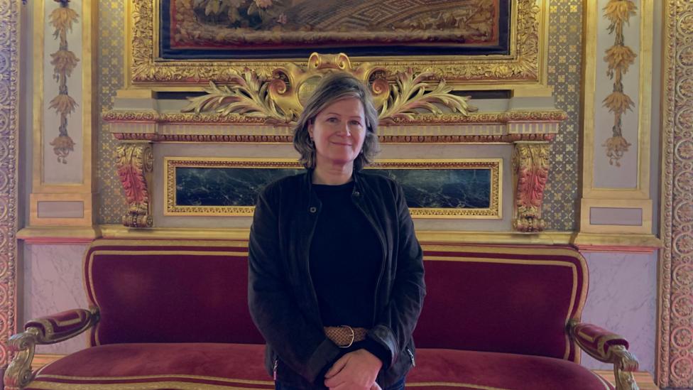 A woman with a dark top and cardigan and greying hair stands in front of an ornate setting with gilded decorations on the wall
