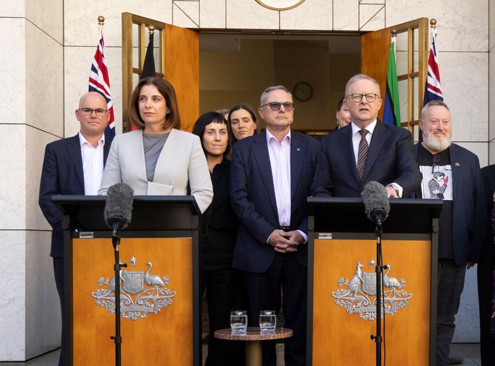 The two politicians stand at podiums in front of a group of parents with the Australian flag behind them and open doors leading onto the terrace of PArliament House in Canberra.
