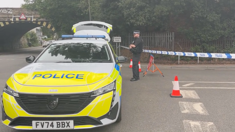 A police car parked. There is a police officer to the right of it and police tape with a road closure sign.