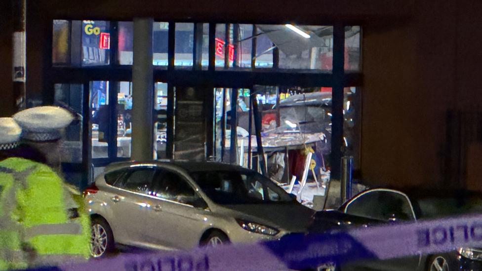 Supermarketning shisha eshigi yonida turgan kumushrang mashina oldida politsiya xodimi turibdi.