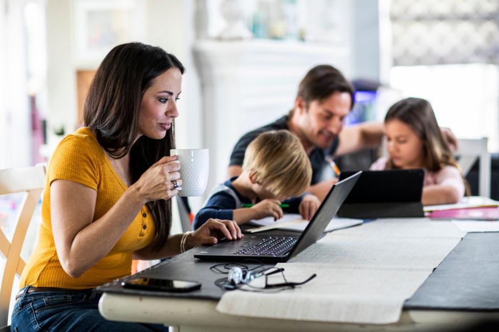 A woman works on a laptop at a dining table while holding a white mug. In the background, two children sit at the same table doing schoolwork, with one child writing on paper and another using a tablet. A man is sat with the children. A smartphone and a pair of eyeglasses are on the table. The scene takes place in a bright, modern home and illustrates working and learning from home simultaneously.