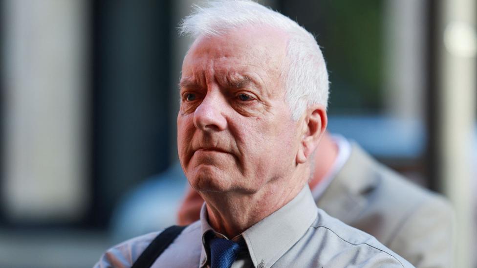 A man in his 70s, with white hair and wearing a light grey shirt with a blue tie, looks past the camera with a blank expression. 