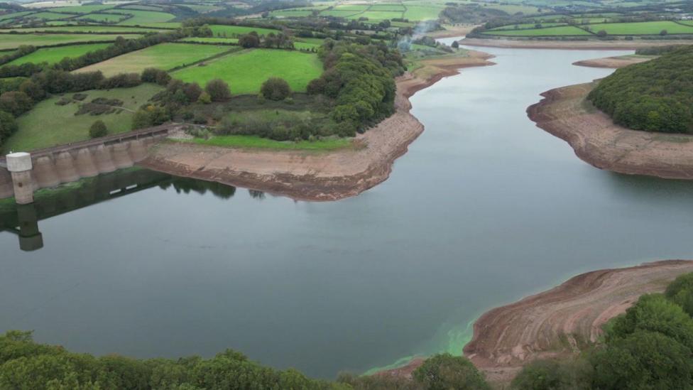 The image shows an aerial view of a reservoir surrounded by patchwork farmland and wooded hills, with large areas of exposed brown shoreline visible around the water's edge. A dam structure can be seen on the left side of the reservoir.