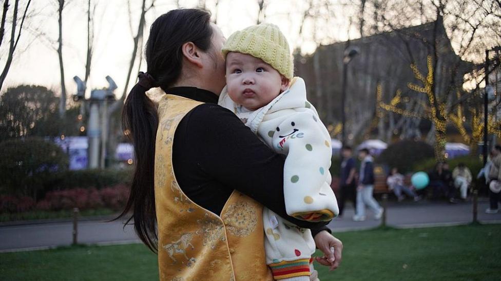 Oqshom vaqti parkda oltin rangli jilet kiygan ayol g‘ubbadon chopon ichiga o‘ralgan chaqaloqni qo‘lida ushlab turibdi