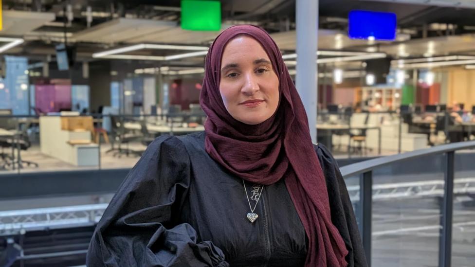 Hannan wearing a burgundy hijab and a black dress. She's stood on a balcony inside the BBC Wales building. There's desks with monitors on them in the background.  