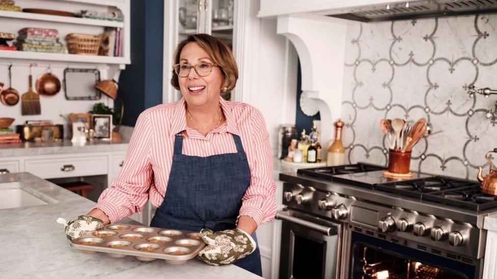 Barbara Costello standing in a kitchen, wearing an apron and holding a muffin tray