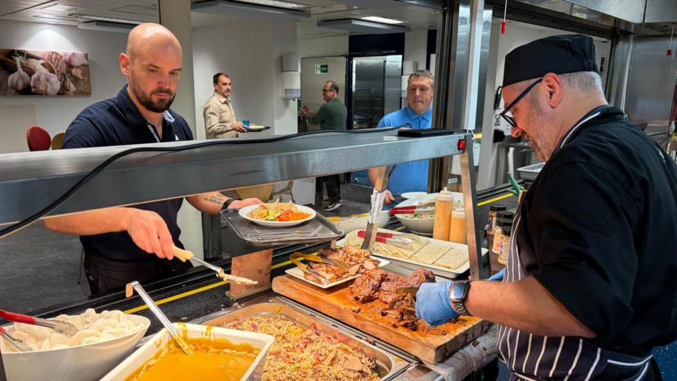 Man serving food in a canteen. He is wearing a black T-shirt and hat and has blue catering gloves on. He is cutting meat. Another man with a bald head and beard is serving himself from another dish