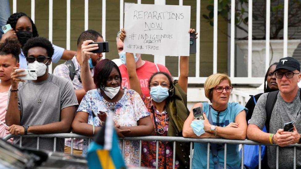 Ayol Britaniya shahzodasi Uilyam va Kembrik gertsogi Ketrinning kelishini kutib turgan paytda norozilik belgisi sifatida plakat ushlab turadi, 2022-yil 25-mart, Karib bo‘ylab safarning yettinchi kuni, Bagama orollari, Nassau