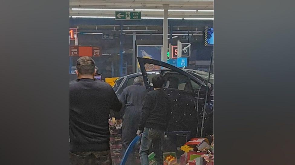 Supermarket ichidagi mashina va uning yonida turgan uch kishining orqa ko‘rinishi.