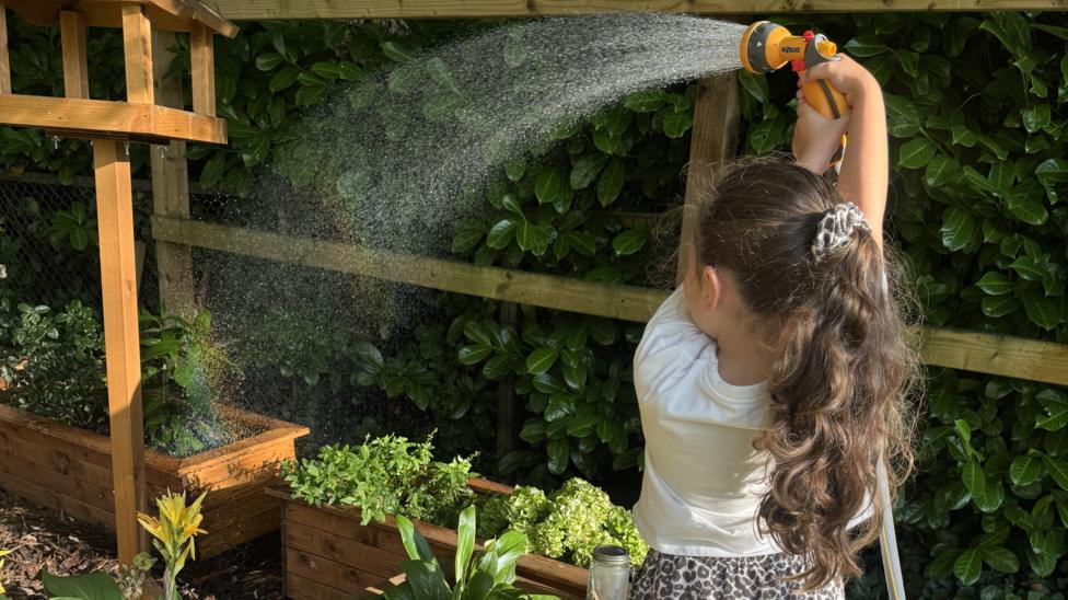 Child standing in a garden in the foreground, holding a hose spray nozzle above their head and watering raised wooden planters. Water arcs from the nozzle across the planters, which contain various flowers and leafy plants. A wooden bird-table-style structure stands beside the planters. A wooden fence and dense hedging form the background.