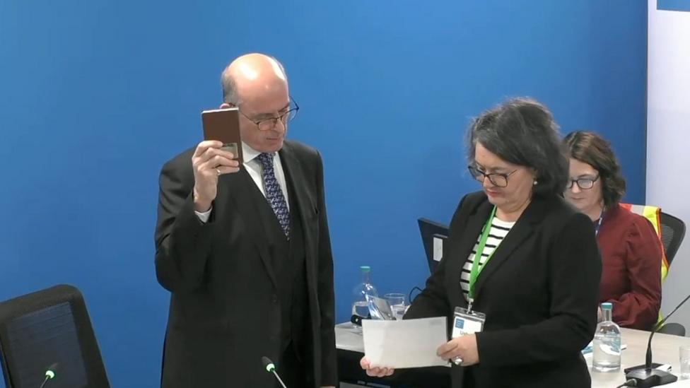 Andrew Coles, wearing a dark suit and tie, holds a Bible in his right hand and is presented with the oath on a piece of paper by the court usher