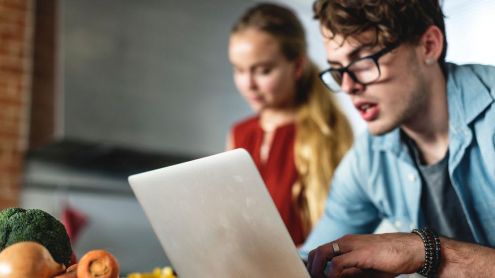 A man looks at a laptop with a woman cooking behind him and some vegetables on a work surface behind the laptop.