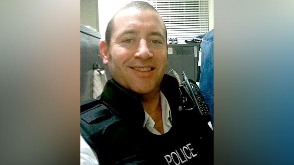 A selfie taken by David Carrick of him sitting in an office with a filing cabinet behind him. He is smiling and has a walkie-talkie strapped to his chest.