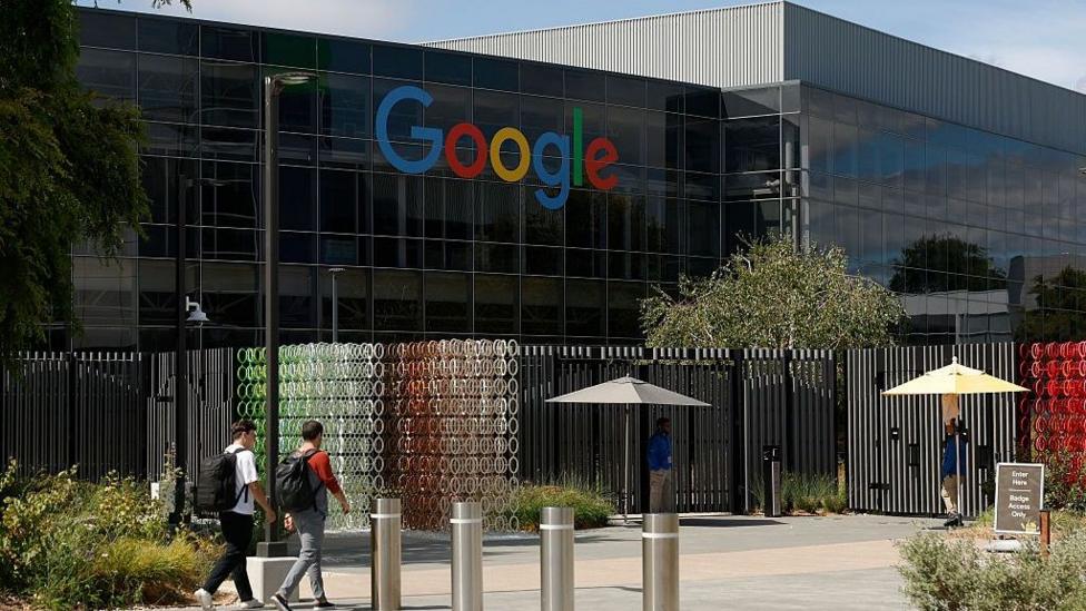 Two unidentified men stroll towards a huge building with the Google logo across its windows.