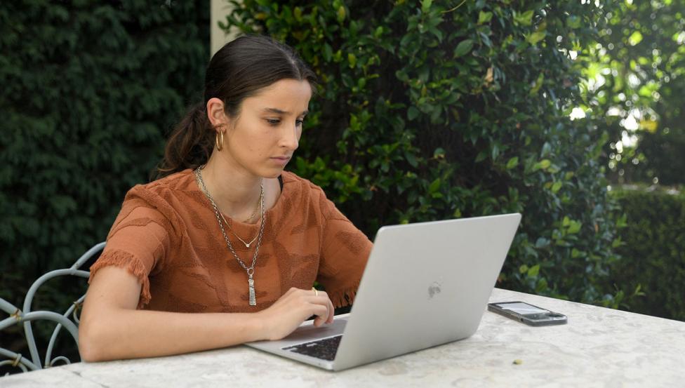 Chanel Contos sits at a table outdoors with a laptop and a phone on the table in front of her while wearing an orange top and necklace, in Sydney in 2022.