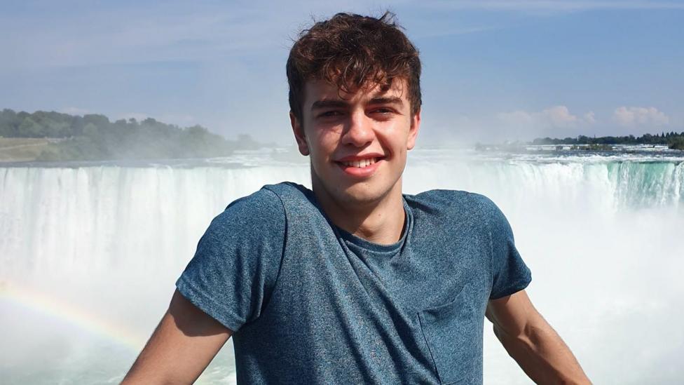 A young man with short brown hair and brown eyes smiling as he stands in front of a waterfall.