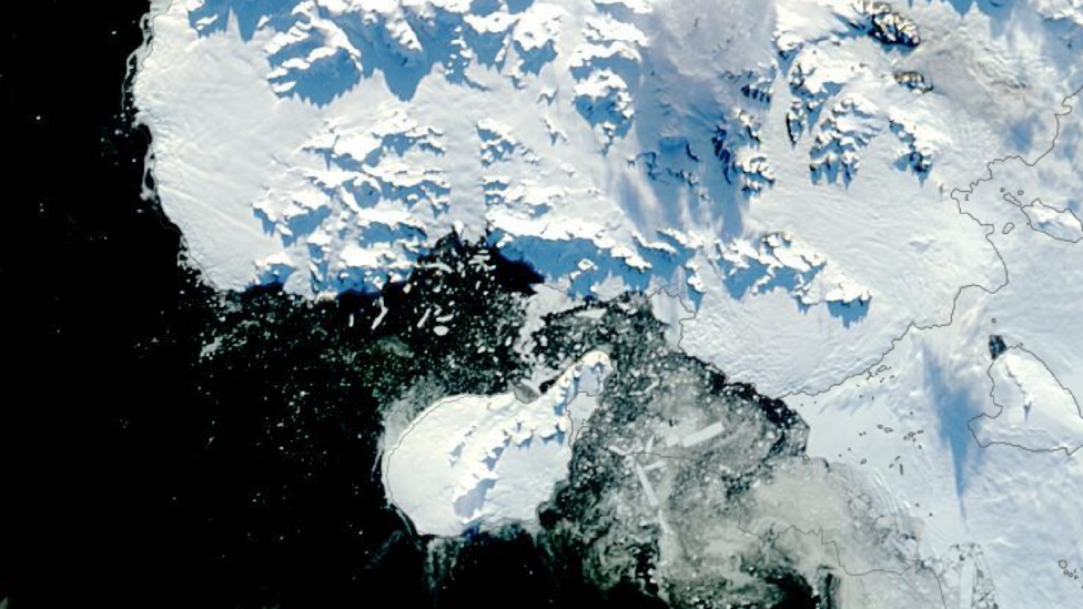 Satellite image of Antarctica. To the left of the image is the ocean, which is black. A small white landmass, which is Rothschild Island, is at the centre bottom of the image. There is a large landmass to the right of the image, which is Alexander Island. The Amos Channel is a stretch of water between the northern tip of Rothschild Island and Alexander Island. The water of the Channel is black with broken ice in it.