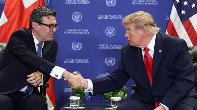 Endi Bernham va Donald Tramp kichik stolning ikki tomonida stullarda o'tirishgan. Ular bir-biriga intilib, qo'l siqishmoqda. Ikkalasi ham to'q rang kostyum kiygan: Bernham ko'k, Tramp esa qizil galstuk taqgan. Ularning orqasida 'BMT Bosh Assambleyasi' so'zlari yozilgan fon ko'rinib turibdi.