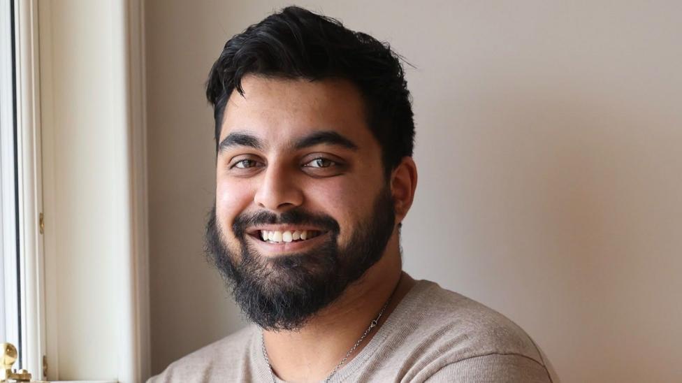 A young man with a beard poses for a portrait. He's next to a window and the light from outside provides a soft, even illumination. He looks relaxed as he smiles at the viewer.