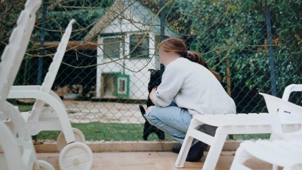 Jessica is kneeling down behind some sun loungers stroking a black dog behind a wire fence. She is wearing blue jeans and a white jumper. The profile is blurred out.
