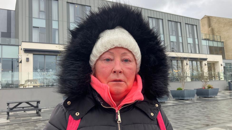 A woman with a black coat, hood up, wearing a white hat looks at the camera. She has on a pink backpack and stands on a grey-stone street in front of a large grey hotel building