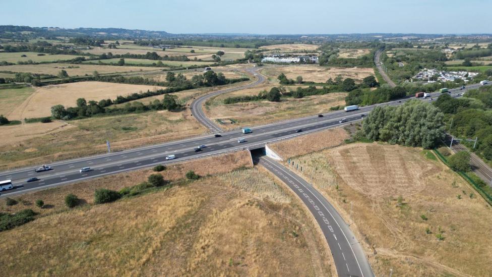 An aerial image of a road stretching beneath another road. It looks very new and there are fields and houses in the distance.