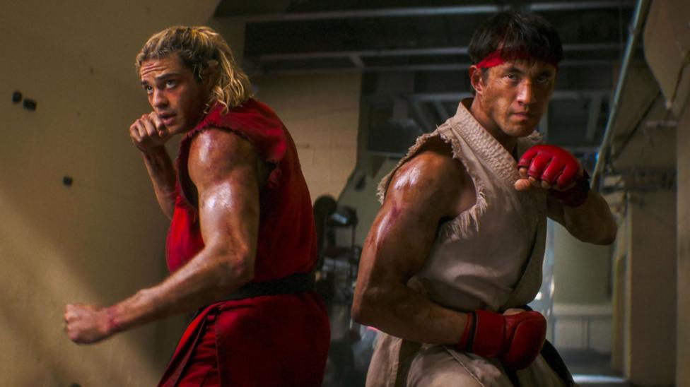 Two men, one with long blonde hair dressed in a red, sleeveless gi (martial arts uniform) and the other in a matching white garment, stand back-to-back, fists pulled back in fighting poses, as they prepare to face an unseen opponent off-camera. Both appear to be dirty and sweating, suggesting they are in the middle of an ongoing battle.