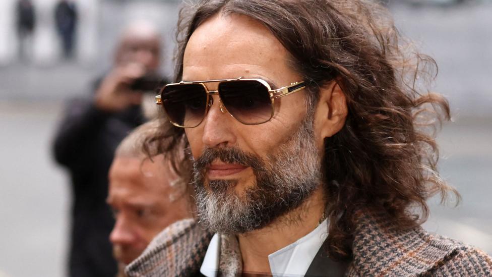 Rassel Brand chorshanba kuni quyoshdan saqlaydigan ko'zoynak va tweed kurtakda sudga kelmoqda