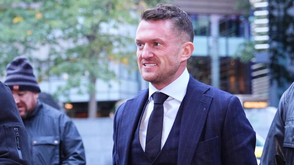 Tommy Robinson sudga borayotgan paytda. U tabassum qilmoqda, yuzida biroz qisiq ifoda bor, to'q ko'k rangli chiziqli galstuk, jilet va kurtka hamda oq ko'ylak kiygan.