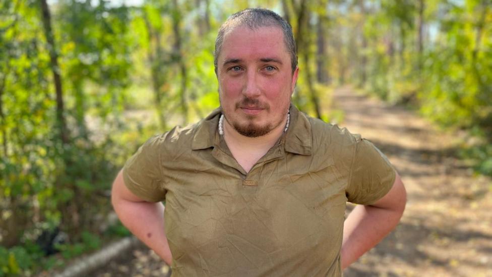 A man in a khaki green polo-shirt stands with his hands on his hips in a sunny forest 