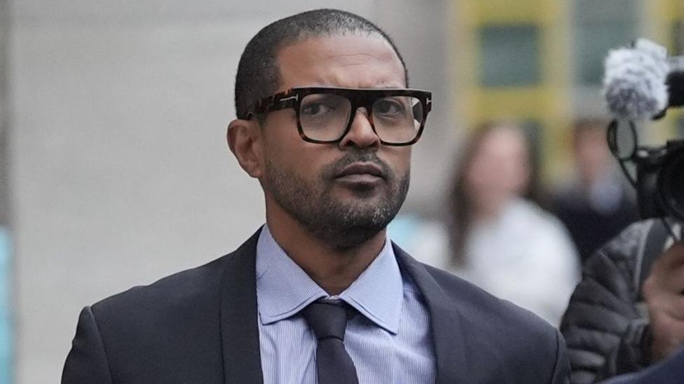Noel Clarke sudga kulrang kostyum, galstuk va ko'zoynak bilan kelmoqda