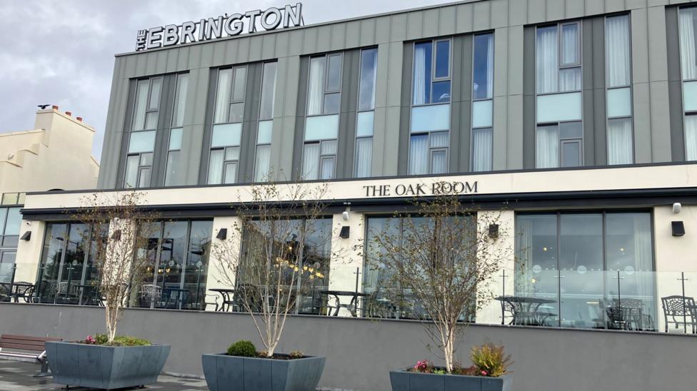 A hotel building has the words The Ebrington on its roof. It is grey and white and sits in front of three, small leafless trees.