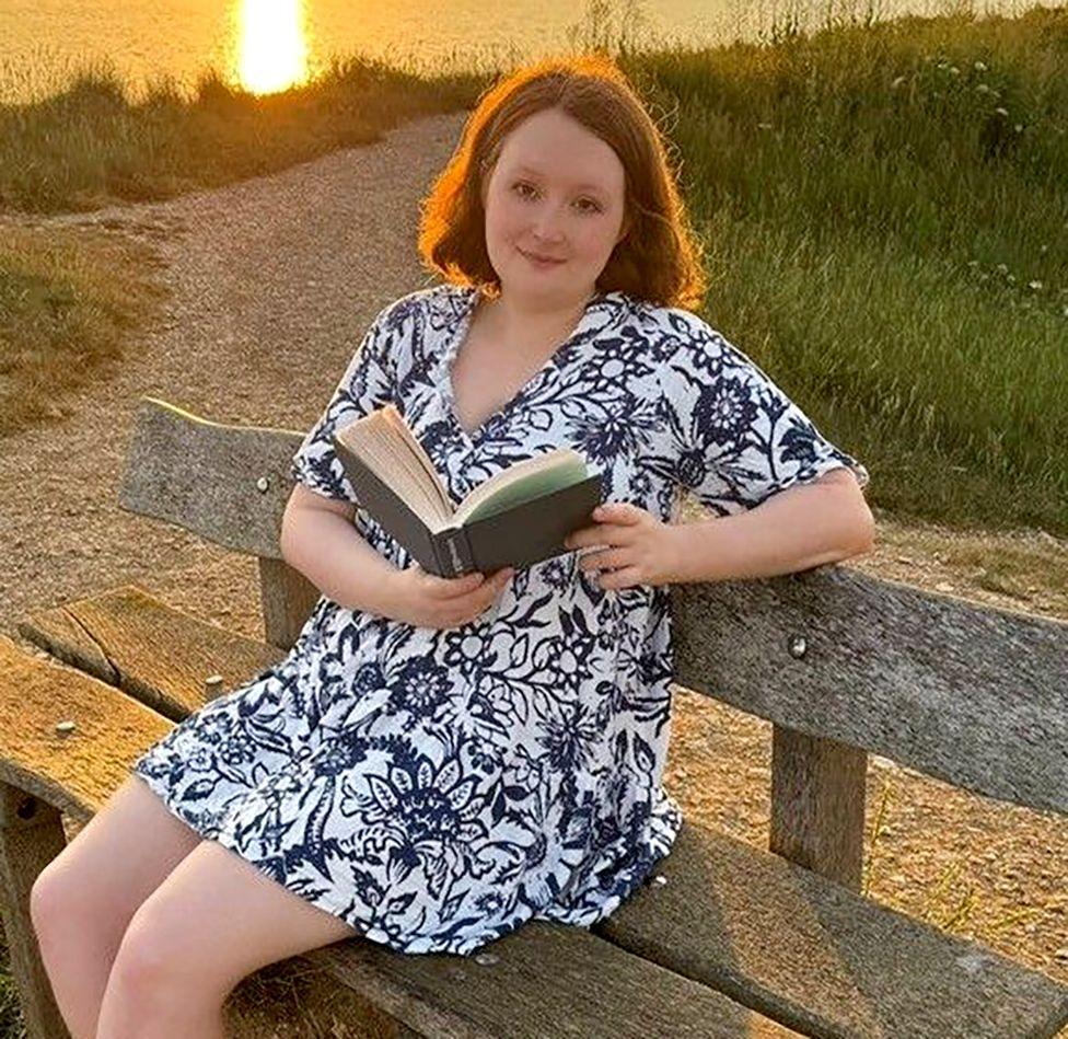 Julia sitting on a bench with an open book beside a lake at sunset.