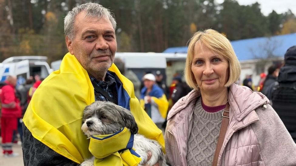 Soçlariga kul rang kirgan Vyaçeslav, yelkasiga Ukraina bayroğini yopinib, qöliga kiçkina itni uşlab turibdi. U sariq soçli bob kesimli rafiqi Olga yonida turibdi. İkkalasi ham kamerga qarayapti.