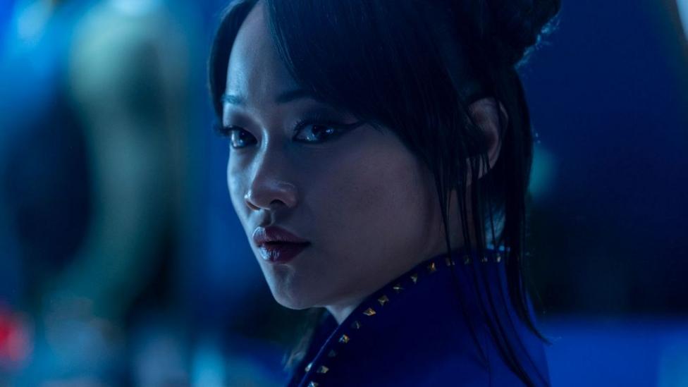 A close up shows Callina Liang as Chun-Li, her hair tied half-up, looking sideways at an object or person of interest off-screen. The shot has a blue hue, giving it a moody, threatening feel.